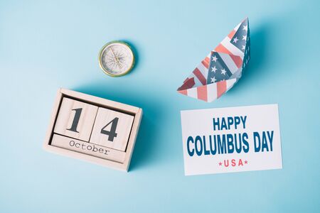 top view of calendar with October 14 date near paper boat with American flag pattern, compass and card with happy Columbus Day inscription on blue background の写真素材