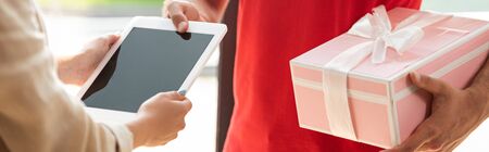 panoramic shot of delivery man holding pink present and giving digital tablet to woman