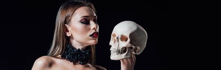 panoramic shot of scary vampire girl in black gothic dress looking at human skull isolated on blackの写真素材