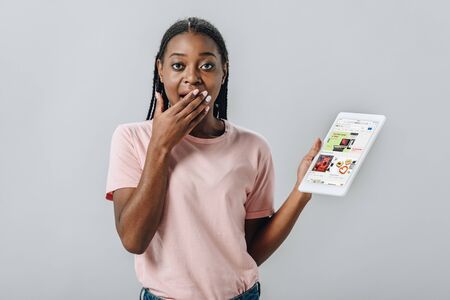 KYIV, UKRAINE - AUGUST 2, 2019: African American woman holding digital tablet with ebay website and covering mouth with hand isolated on greyのeditorial素材