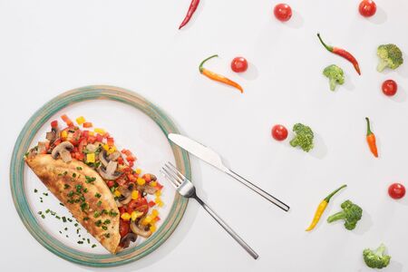 top view of plate with homemade wrapped omelet with vegetables on white table with chili peppers,...