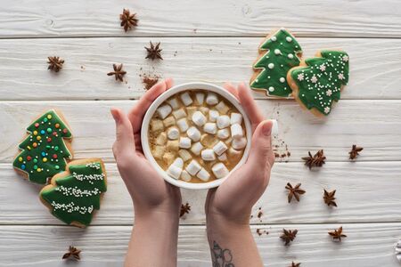 cropped view of woman holding mug with cacao and marshmallows at white wooden table with anise and...