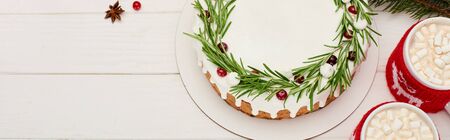 top view of christmas pie with icing, rosemary and cranberries on white wooden table with cups of cocoaの写真素材