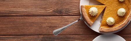 panoramic shot of delicious pumpkin pie with whipped cream on plate with spatula on brown wooden tableの写真素材