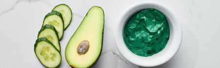 panoramic shot of cucumber slices, half of avocado and green face mask on marble surface