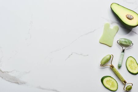 top view of facial spatula and rollers near avocado and cucumber on marble surfaceの写真素材