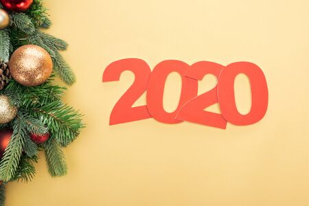 top view of paper 2020 numbers near christmas tree branch with baubles on yellow background