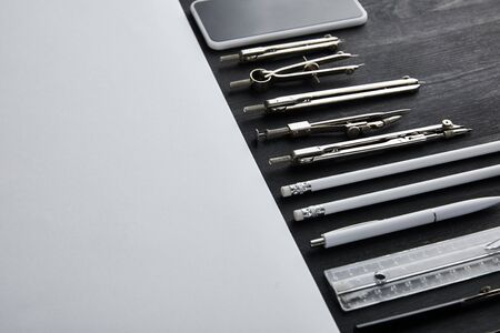 white paper with copy space and compasses, ruler, pencils, smartphone, pen on black and wooden table