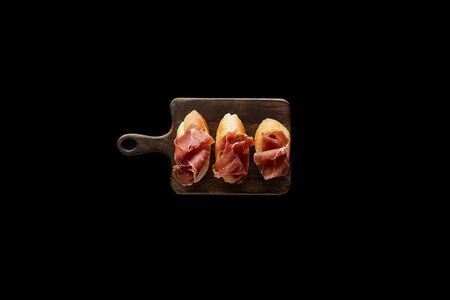 top view of sliced prosciutto on baguette on wooden cutting board isolated on blackの写真素材