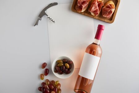 top view of bottle with rose wine near grape, corkscrew, olives, blank paper and sliced prosciutto on baguette on white backgroundの写真素材