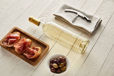 bottle with white wine near prosciutto on baguette, olives and corkscrew on napkin on white wooden...