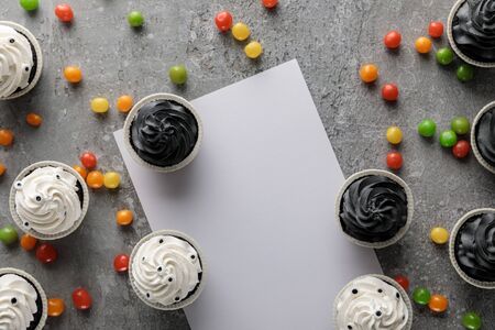 top view of delicious Halloween cupcakes with scattered colorful bonbons near white blank paper on concrete grey surfaceの写真素材