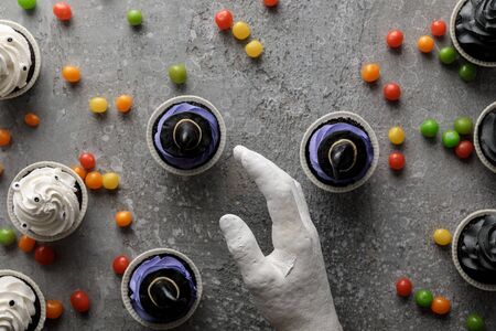 top view of decorative hand near delicious Halloween cupcakes with scattered colorful bonbons on concrete grey surfaceの写真素材