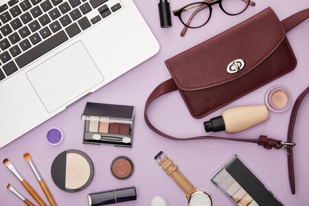 top view of bag and decorative cosmetics near laptop isolated on violetの写真素材