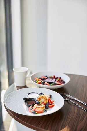 traditional Ukrainian varenyky and syrniki with berries served in white plate with sauce near cutlery and cup on wooden table in restaurantの写真素材