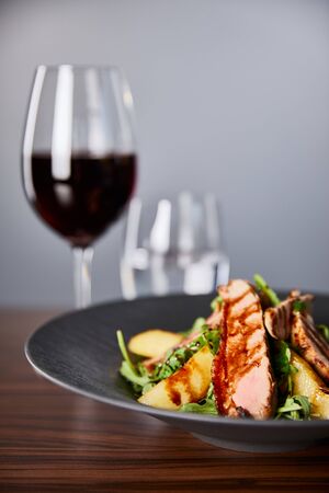 selective focus of delicious warm salad with arugula, potato and meat in black plate on wooden table near red wine, water on grey backgroundの写真素材