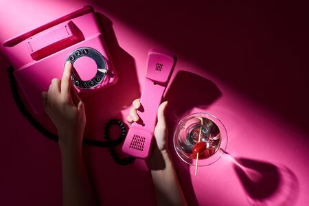 Cropped view of woman using vintage telephone beside cocktail on pink backgroundの写真素材