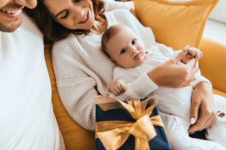 cropped view of smiling man sitting near happy wife holding adorable baby near gift box with golden ribbonの写真素材