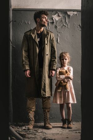 handsome man standing near kid with teddy bear near weathered wall, post apocalyptic conceptの写真素材
