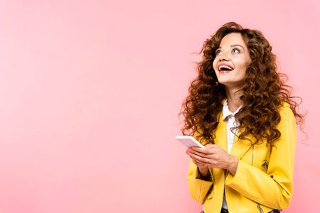 excited curly girl using smartphone, isolated on pinkの写真素材