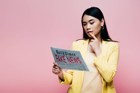 thoughtful asian woman in yellow outfit reading newspaper with fake news isolated on pinkの写真素材