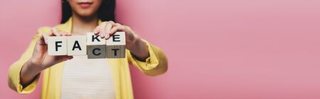 cropped view of asian woman holding wooden cubes with fake and fact lettering isolated on pink, panoramic shotの写真素材