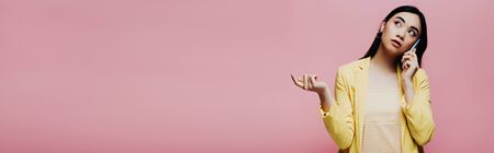 dreamy asian woman in yellow outfit talking on smartphone isolated on pink, panoramic shotの写真素材