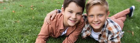 panoramic shot of two happy brothers smiling at camera while lying on grass