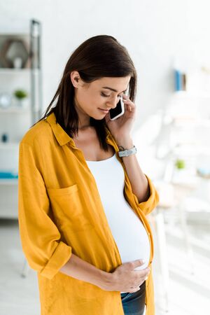 beautiful pregnant freelancer in yellow shirt talking on smartphone in home officeの写真素材