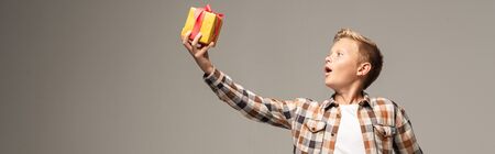 panoramic shot of amazed boy holding gift box in outstretched hand isolated on grey