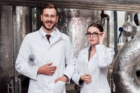 happy scientist holding blueprints near beautiful coworker with digital tablet