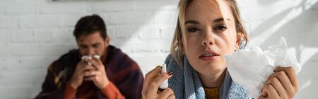 sick woman holding nasal spray and napkins in bedroom with man behind