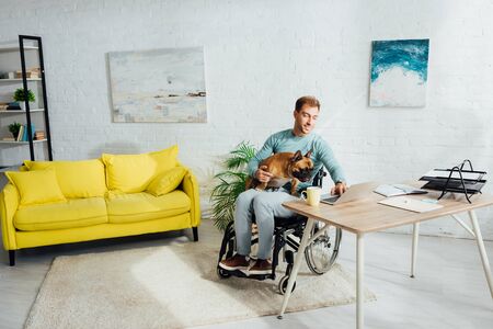 Disabled man holding french bulldog on knees and working on laptop at home