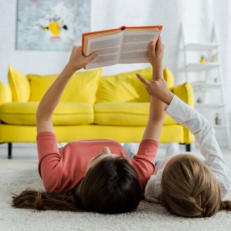 kid pointing with finger at book while lying on carpet with babysitter