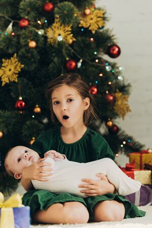 amused child looking at camera while sitting near christmas tree and holding cute little sisterの写真素材