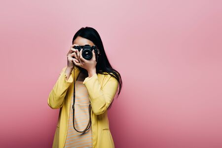 asian woman in yellow outfit taking picture on digital camera isolated on pinkの写真素材