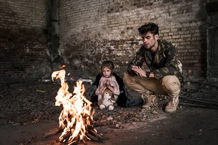 selective focus of man and child sitting near bonfire, post apocalyptic concept