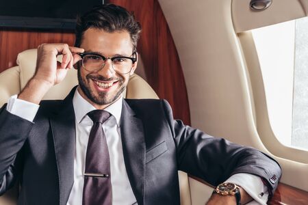 handsome businessman in glasses smiling and looking at camera in private planeの写真素材