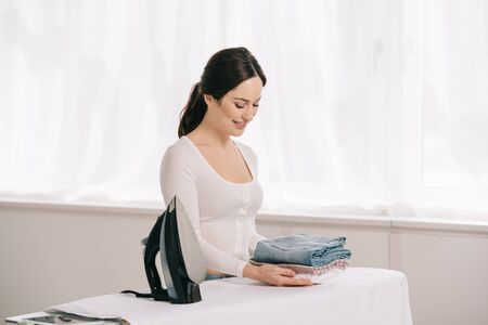 smiling housewife holding ironed clothes while standing near ironing boardの写真素材