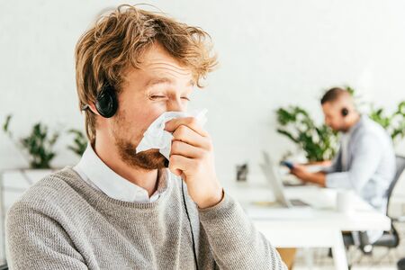 sick bearded broker with closed eyes sneezing in tissue near coworker in officeの写真素材