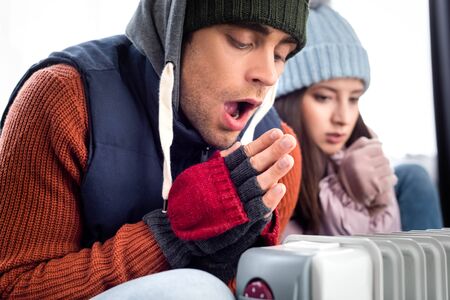 selective focus of girlfriend and boyfriend in winter outfit warming up near heater