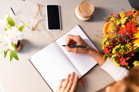 cropped view of woman writing in notepad in cafe with coffee, smartphone and flowersの写真素材