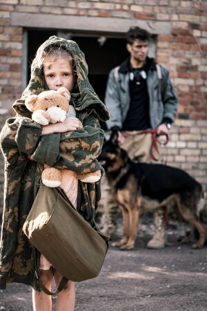 selective focus of kid holding teddy bear near man with german shepherd dog near abandoned building, post apocalyptic conceptの写真素材