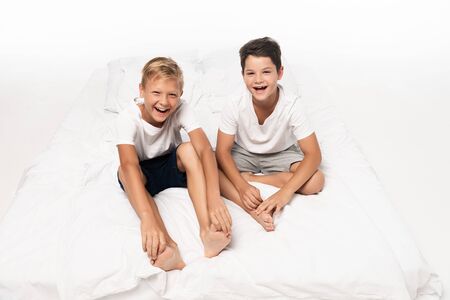 two happy brothers sitting on bed and smiling at camera on white backgroundの写真素材