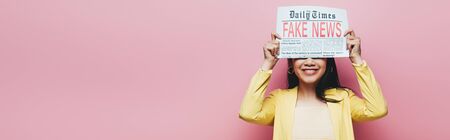 panoramic shot of happy asian woman in yellow outfit holding newspaper with fake news isolated on pinkの写真素材