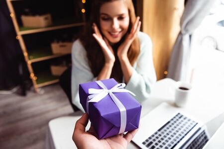 selective focus of surprised woman taking gift in cafe with laptop and cup of coffee