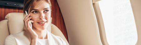 panoramic shot of smiling businesswoman in suit talking on smartphone in private plane