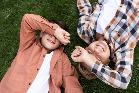 top view of two cheerful brothers covering eyes with hands while lying on green grassの写真素材