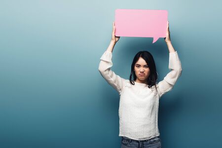 displeased brunette asian woman holding pink speech bubble on blue background