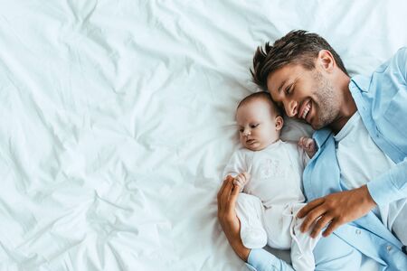 top view of happy father hugging adorable baby while lying on white beddingの写真素材